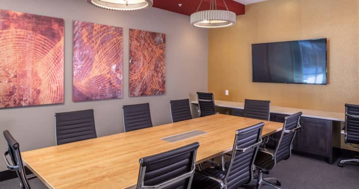 Conference room with long wooden table, black ergonomic chairs, abstract wall art, mounted TV screen, and pendant lighting.