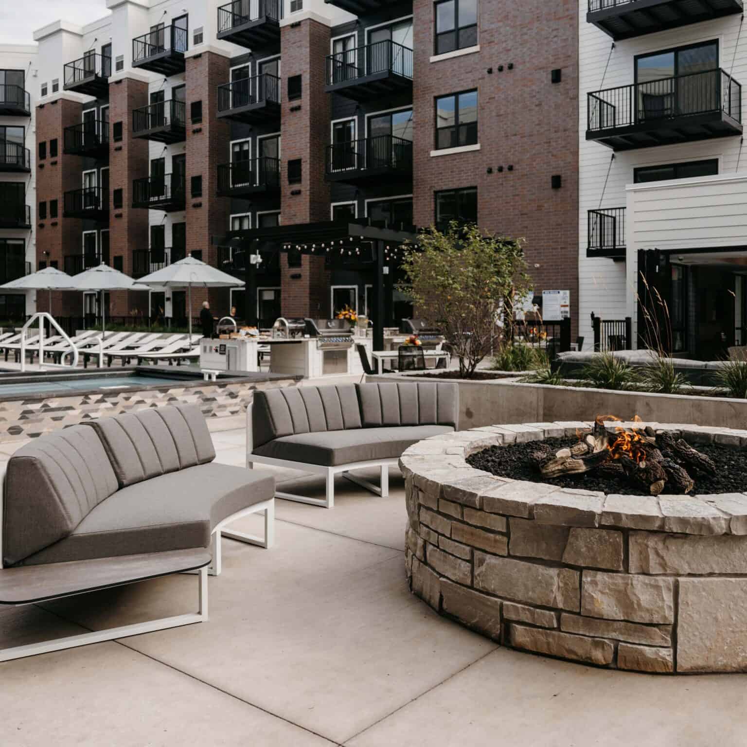 Outdoor courtyard with curved gray sectional seating, round stone fire pit with active fire, and multi-story brick apartment buildings in background.