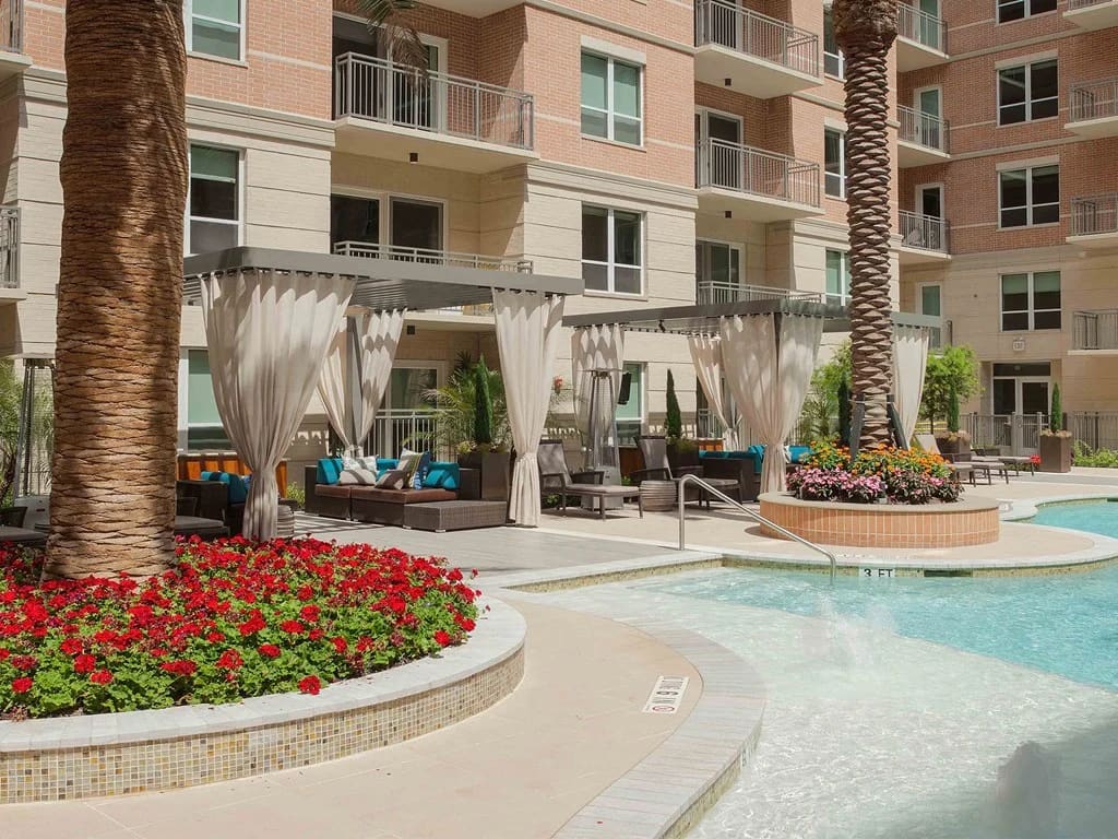 Courtyard pool area with palm trees, red flower beds, white pergolas with lounge seating, and multi-story brick apartment building.