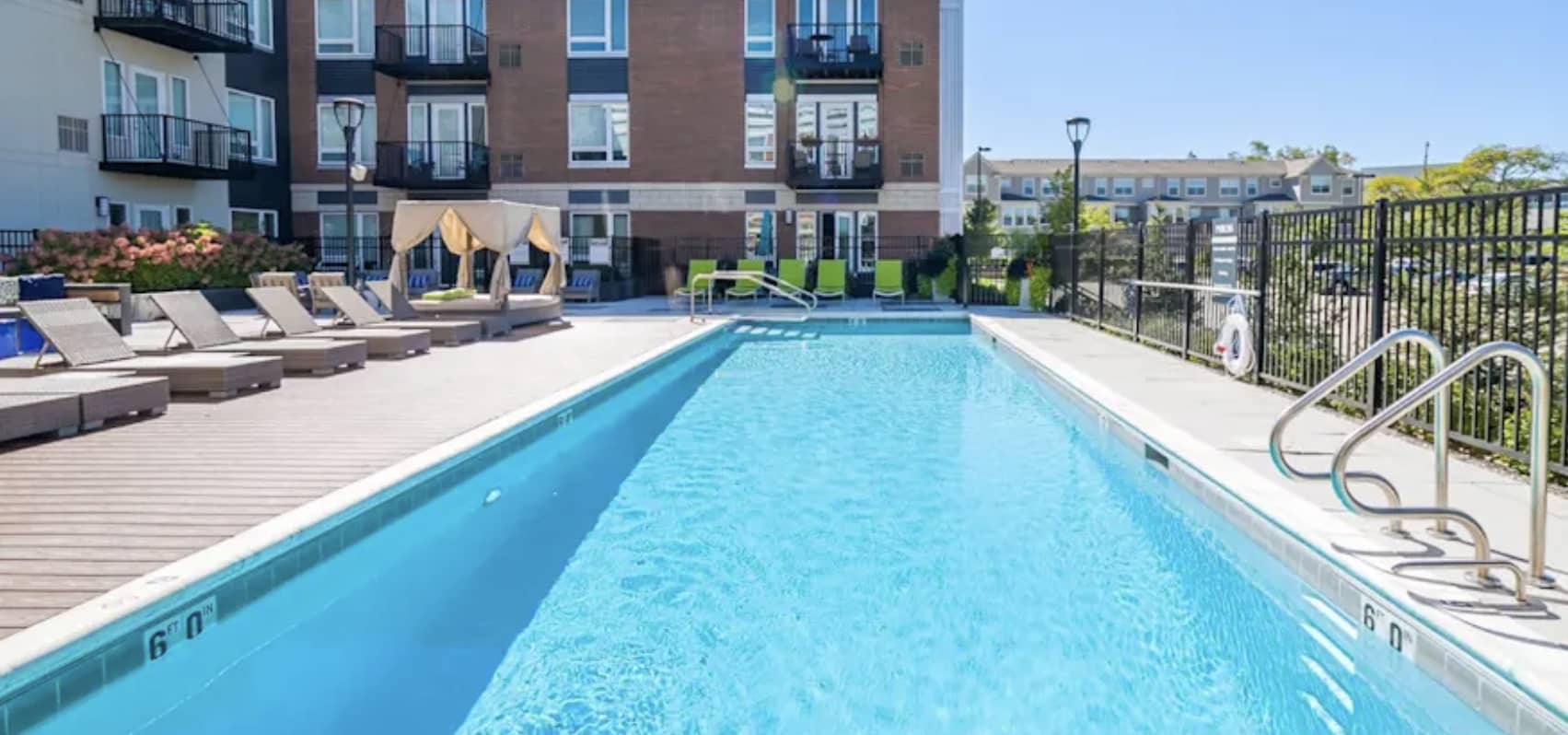 Rectangular outdoor swimming pool with clear blue water, lounge chairs, and brick residential building in background.