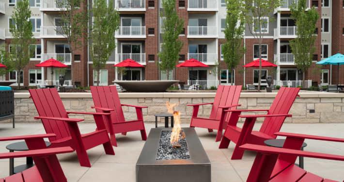 Courtyard with four red Adirondack chairs arranged around a linear fire pit, brick residential buildings with balconies in background.
