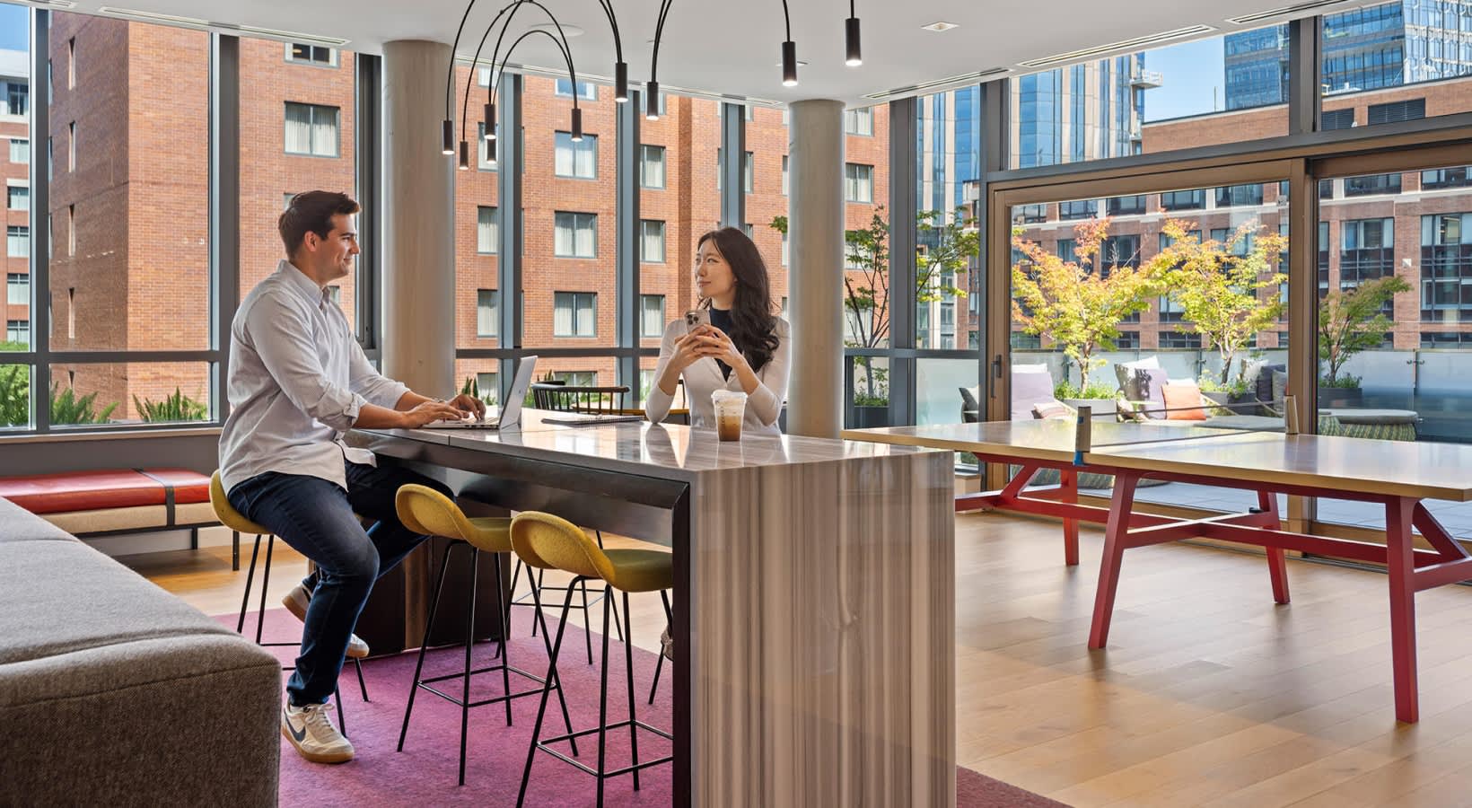 Two people at a high-top table in a bright common room with large windows, pendant lights, and views of brick buildings and green courtyard space.