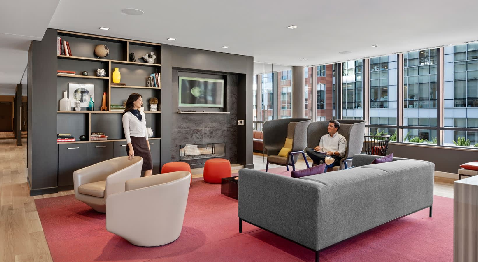 Modern lounge with gray sofa, cream curved chair, red rug, dark shelving unit with TV, and floor-to-ceiling windows overlooking city buildings.