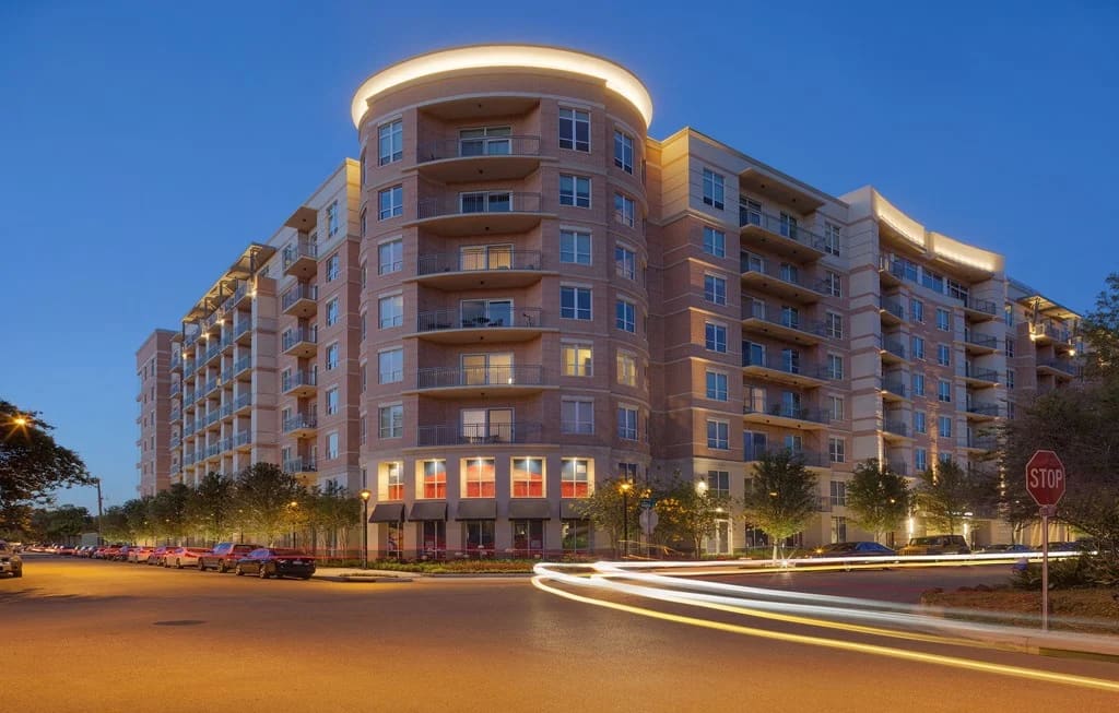 Multi-story brick apartment building with curved corner tower, tiered balconies, and illuminated ground-floor storefronts at dusk.