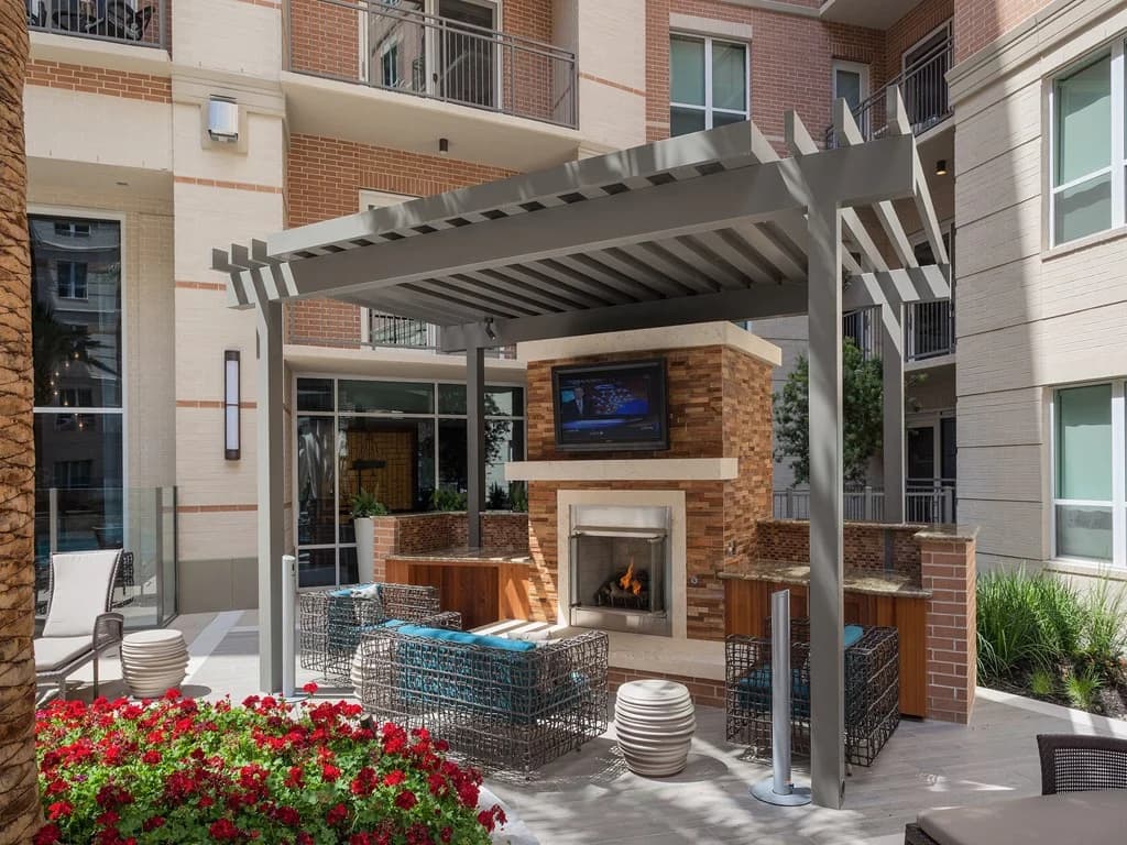 Courtyard with gray pergola, brick fireplace with TV, woven seating, red flowers, and surrounding apartment buildings.