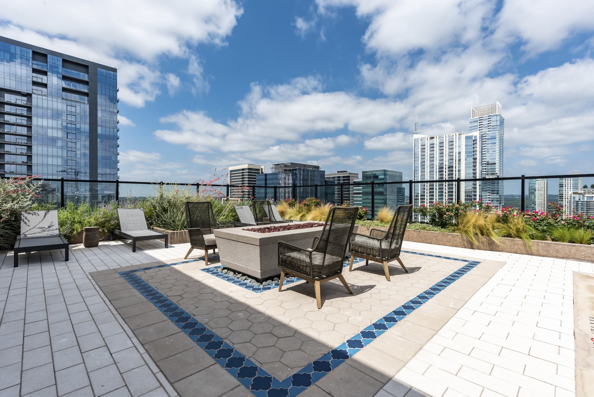 Rooftop terrace with lounge chairs, fire pit, potted plants, and city skyline views under blue sky.