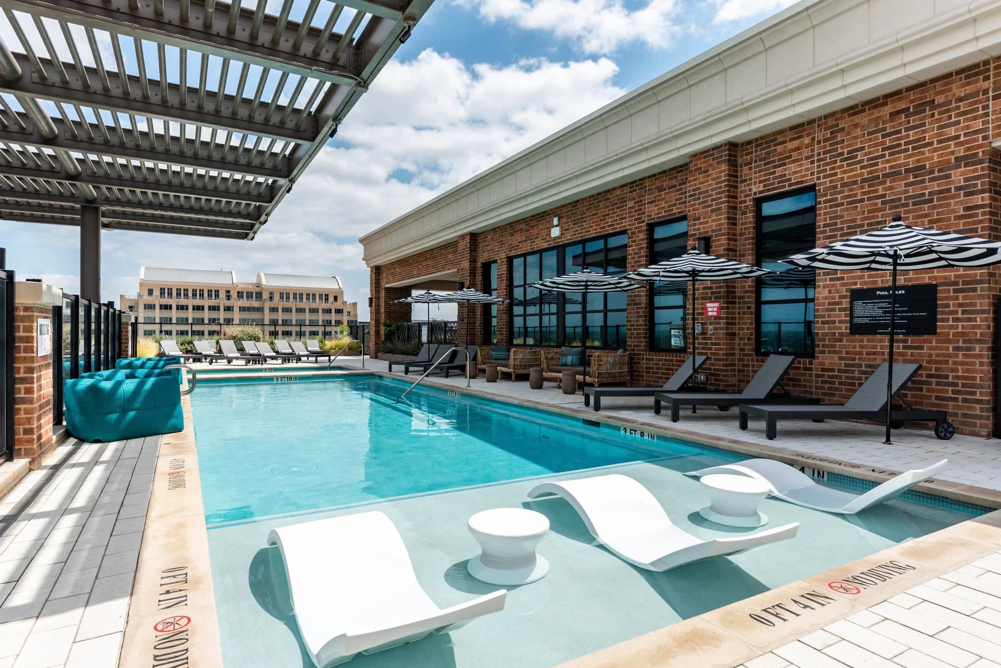 Rooftop pool deck with blue water, white lounge chairs, hot tub, striped umbrellas, and brick building with windows overlooking downtown Austin.