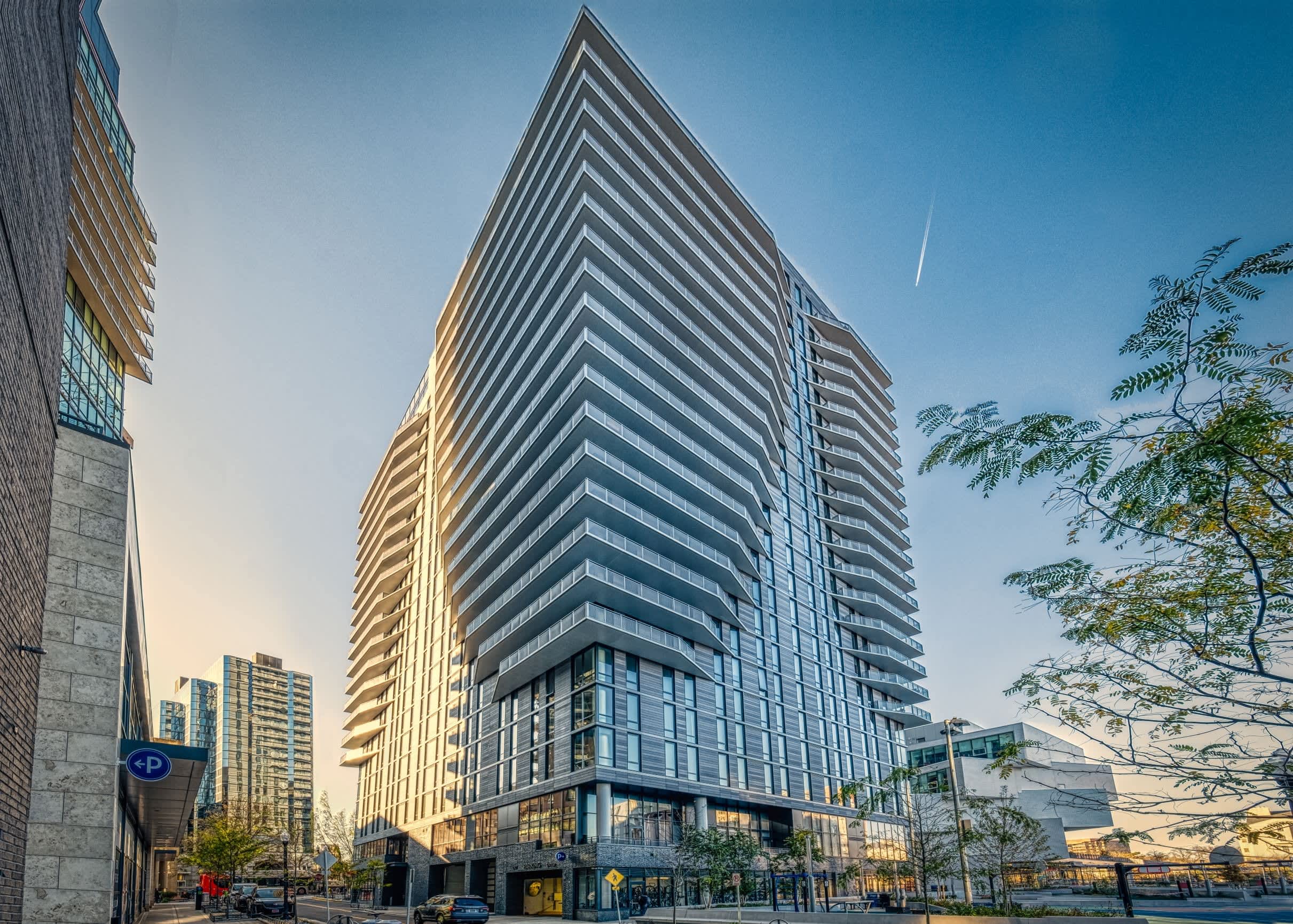 Modern residential high-rise building with horizontal striped balconies, ground-floor retail, and tree-lined street in urban setting.
