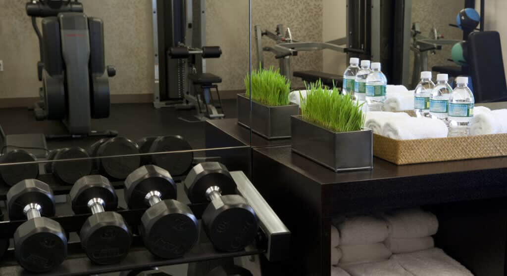 Fitness center with rows of dumbbells in foreground, exercise bikes and equipment in background, black counter with green plants and water bottles, white towels stacked below.