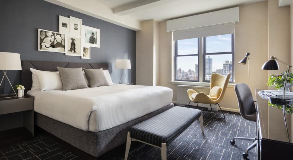 Hotel bedroom with gray accent wall, white bedding, floor-to-ceiling windows overlooking Manhattan skyline, and yellow accent chair.