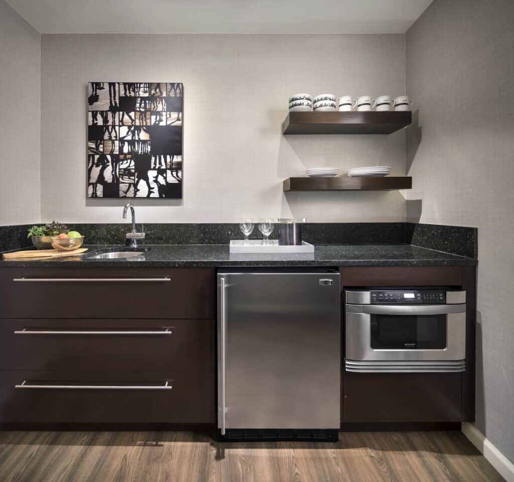 Kitchenette with dark wood cabinets, black granite countertop, stainless steel refrigerator and microwave, open shelving with dishware, and abstract wall art.