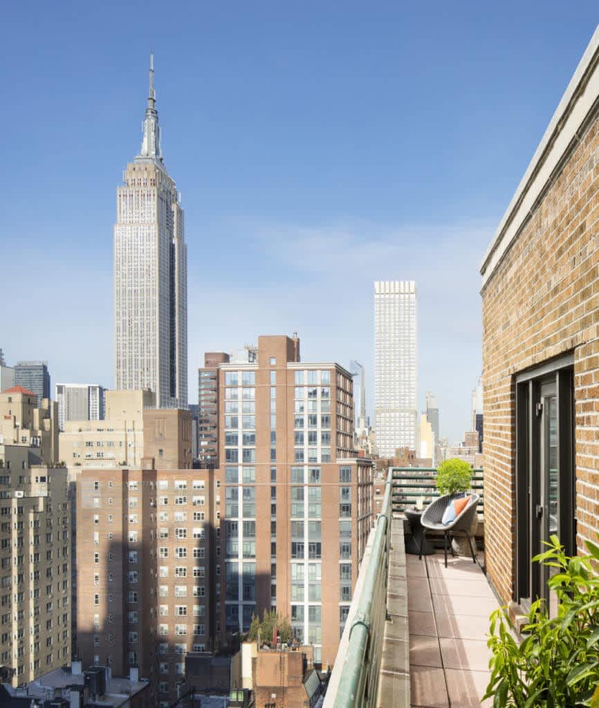Rooftop terrace with lounge chair and potted plants overlooking Manhattan skyline with Empire State Building and surrounding skyscrapers under clear blue sky.