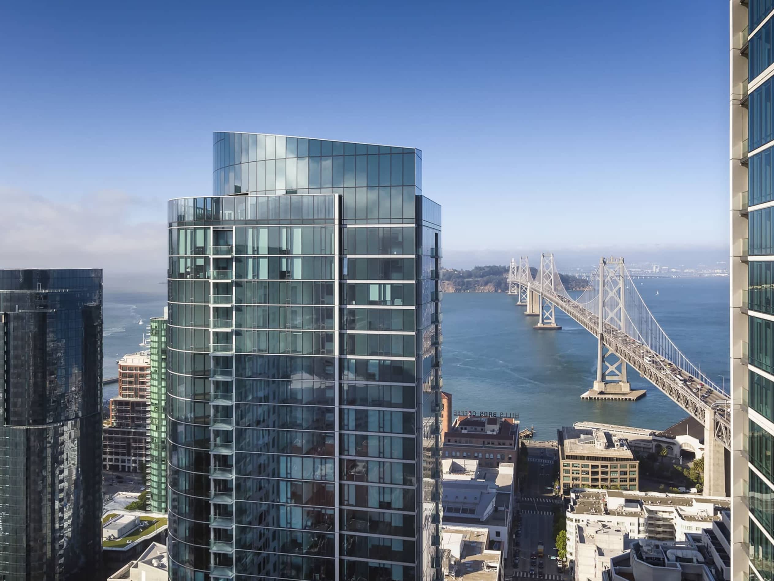 High-rise residential tower with Bay Bridge and San Francisco Bay visible in the background under clear blue sky.