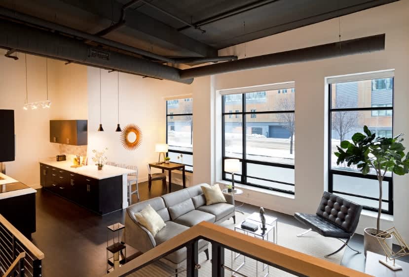Open loft living space with gray sofa, dark kitchen island, pendant lights, and floor-to-ceiling windows overlooking downtown Milwaukee.