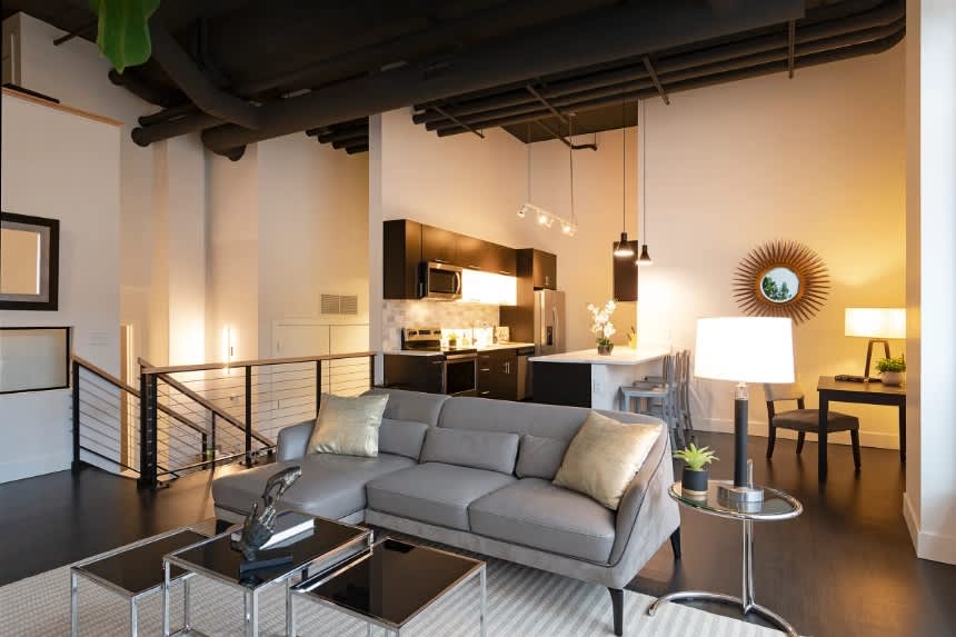 Modern loft living room with gray sofa, exposed black ceiling beams, open kitchen with dark cabinetry, and mezzanine level with metal railing.