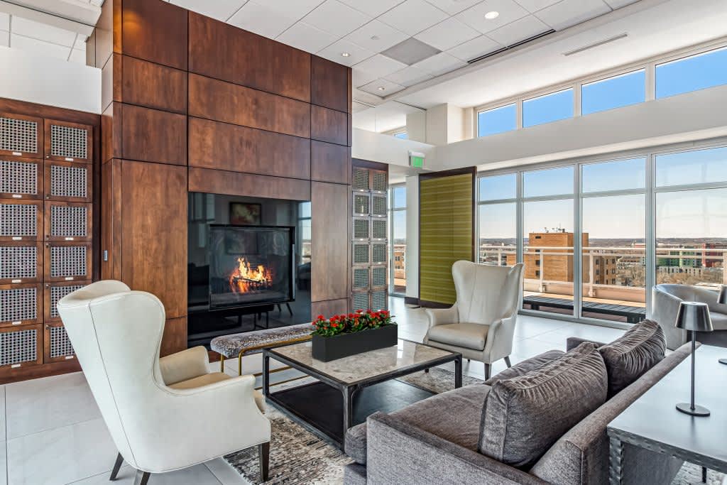 High-rise living room with wood-clad fireplace, gray sofa, white wingback chair, and floor-to-ceiling windows overlooking Madison cityscape.