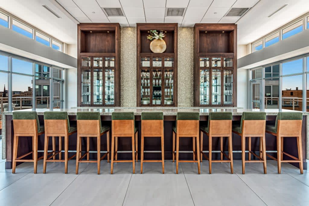 Modern lounge with ten leather and wood bar stools facing built-in wood cabinetry with glass doors, large windows, and city views.