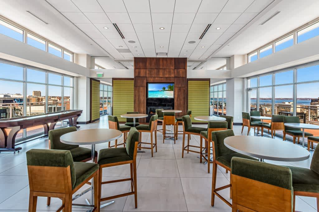 High-rise lounge with round white tables, green upholstered chairs with wood frames, wood accent wall with TV, floor-to-ceiling windows overlooking Madison cityscape.