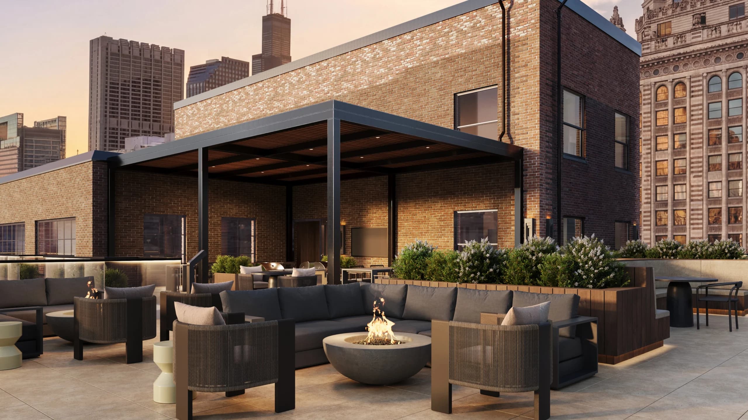 Rooftop terrace with dark gray sectional seating, round fire pit, potted plants, and Chicago skyline in background at sunset.