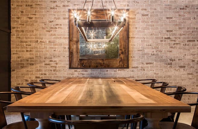 Dining room with long wooden table, black chairs, neon-framed artwork on brick wall, and geometric pendant light fixture.