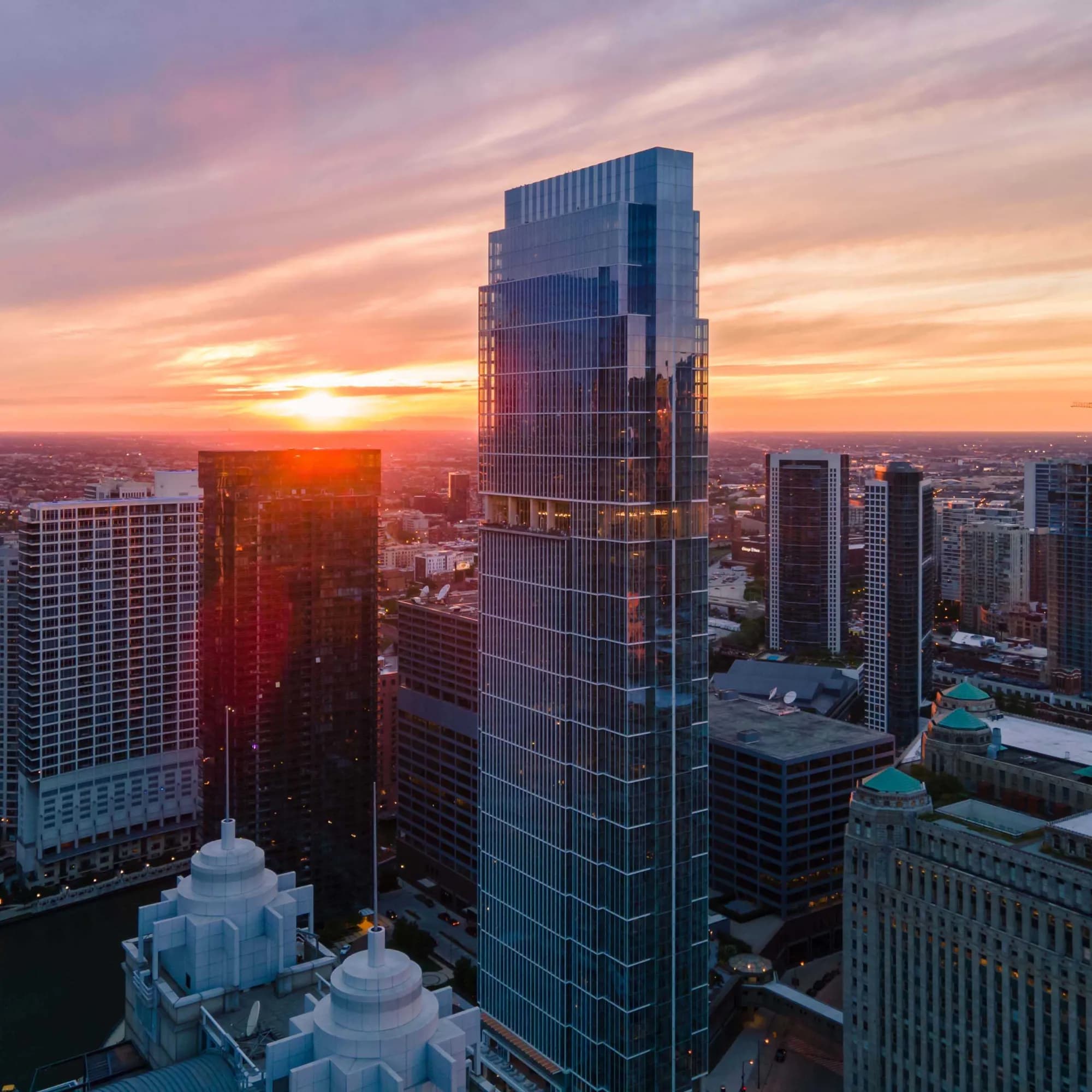 Modern high-rise residential tower at sunset with Chicago skyline, surrounding buildings, and golden sky in background.