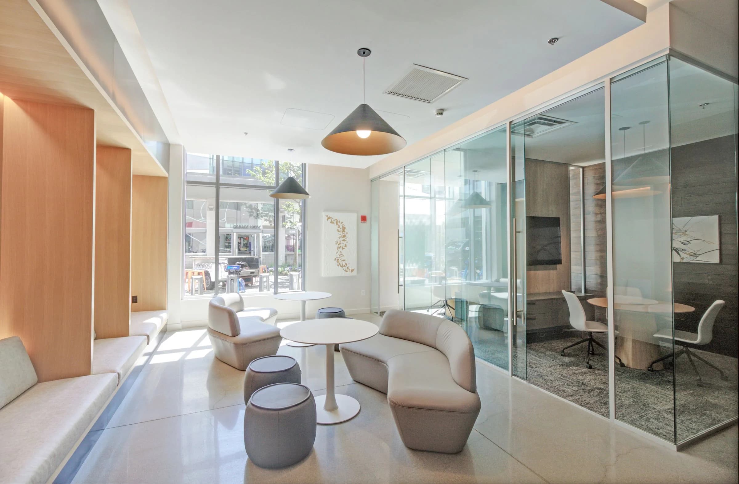 Bright common lounge with cream curved seating, round white table, pendant lights, and glass-enclosed office spaces with street views.