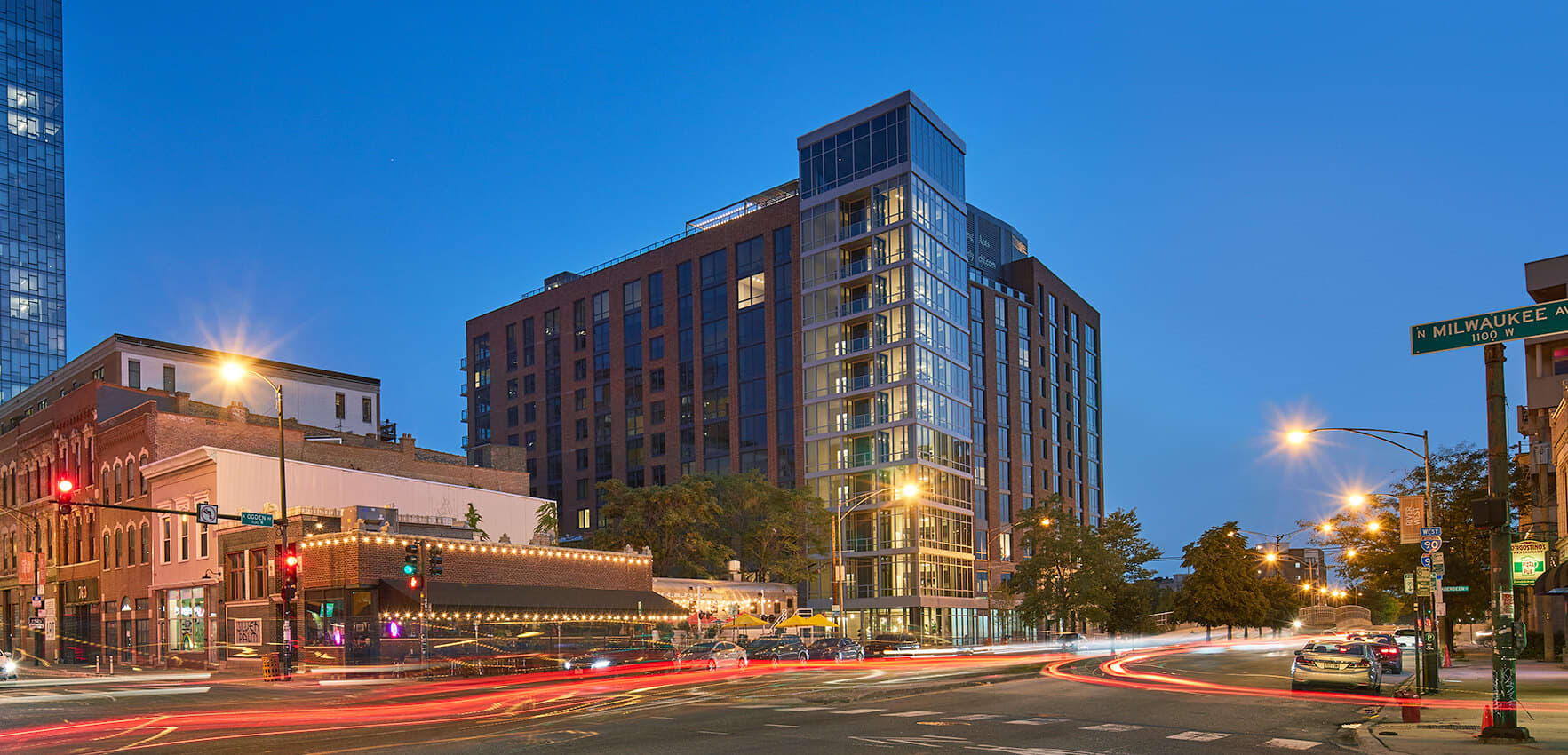 Modern mixed-use building with illuminated windows at dusk on a Chicago street corner, surrounded by historic brick buildings and evening traffic.