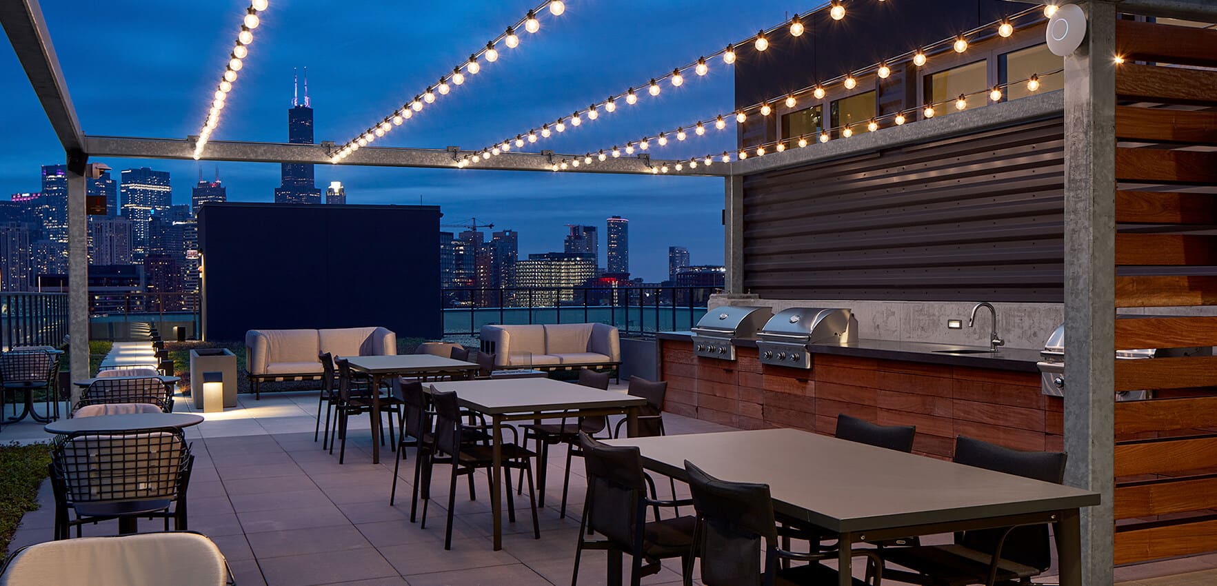 Rooftop terrace at dusk with string lights, dining tables, lounge seating, built-in grill, and Chicago skyline backdrop.
