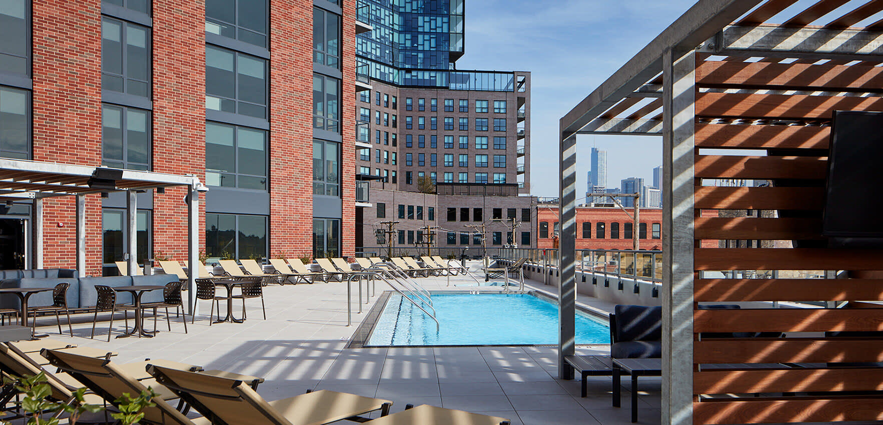 Rooftop pool with lounge chairs, pergola shade structure, and surrounding brick residential buildings in urban setting.