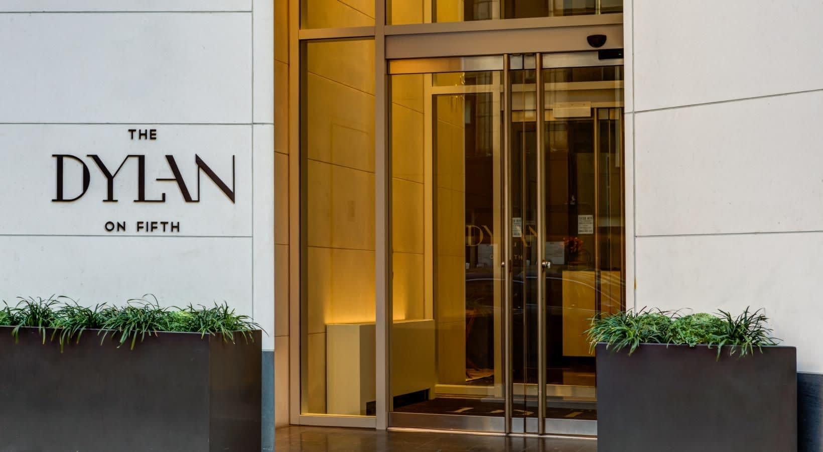 Building entrance with brass glass doors, white facade, and black planters with green plants flanking the entry to The Dylan on Fifth.