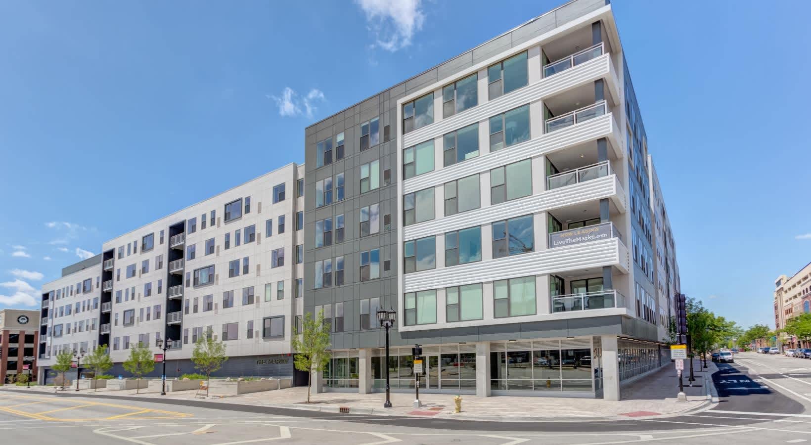 Contemporary multi-story apartment building with white and gray facade, balconies, and ground-floor retail storefronts on a tree-lined street.
