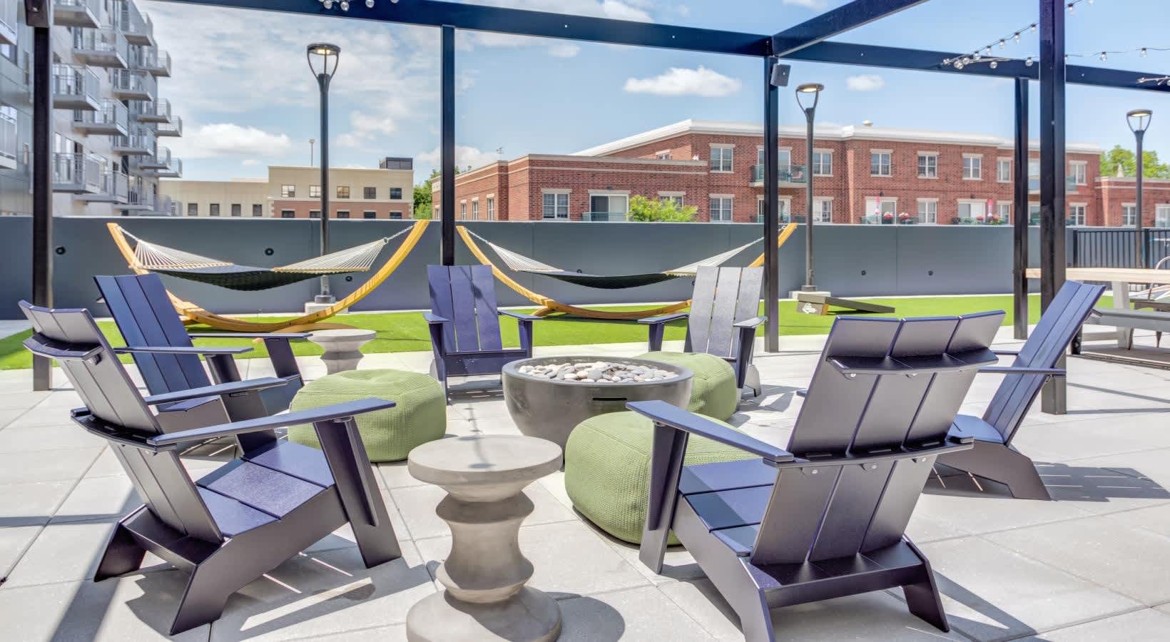 Rooftop terrace with blue Adirondack chairs, fire pit, hammocks, and green lawn surrounded by brick buildings and city skyline.