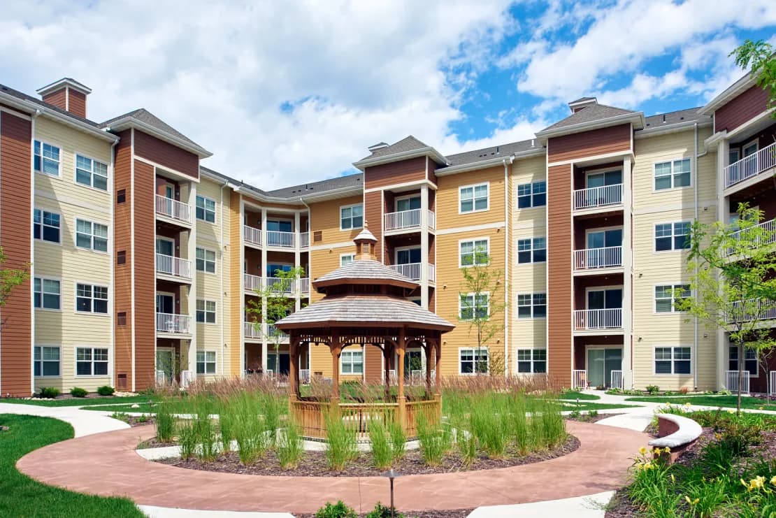 Multi-story apartment complex with red, yellow, and cream siding surrounding a central courtyard with wooden gazebo and landscaping.