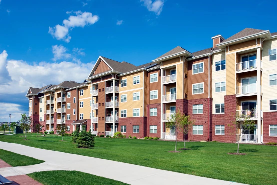 Multi-story apartment complex with brick and tan siding, white balconies, manicured lawn, and concrete walkway under blue sky.