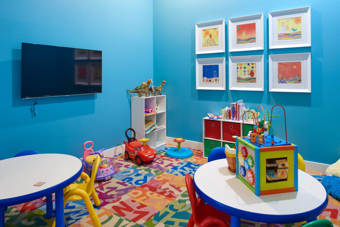 Bright blue playroom with two white activity tables, colorful toys, wall-mounted TV, framed artwork, and storage shelves with books and toys.