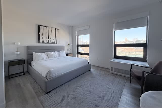 Modern bedroom with gray upholstered bed, white bedding, nightstand, and floor-to-ceiling windows overlooking city buildings.