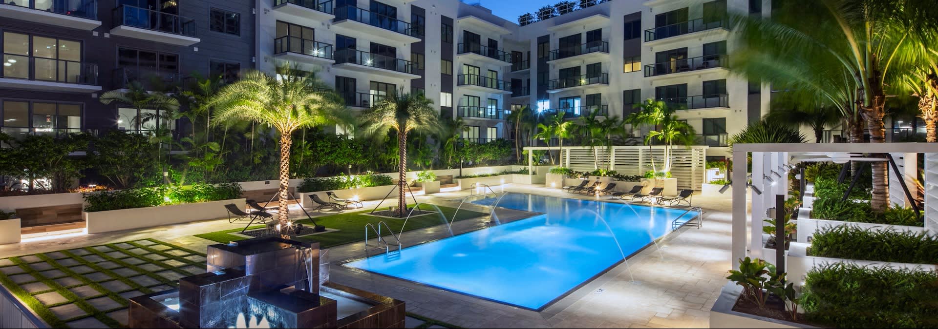Illuminated resort pool with lounge chairs, palm trees, and modern residential buildings at dusk in Miami.