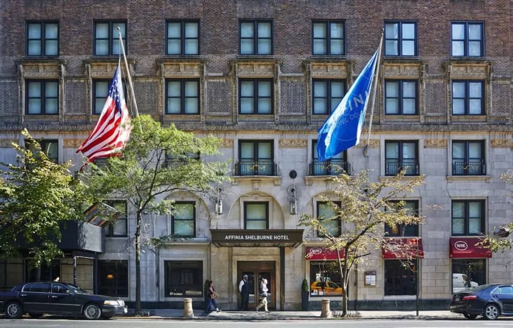 Historic brick and stone hotel facade with arched windows, American and blue flags, green trees, and ground-floor entrance.
