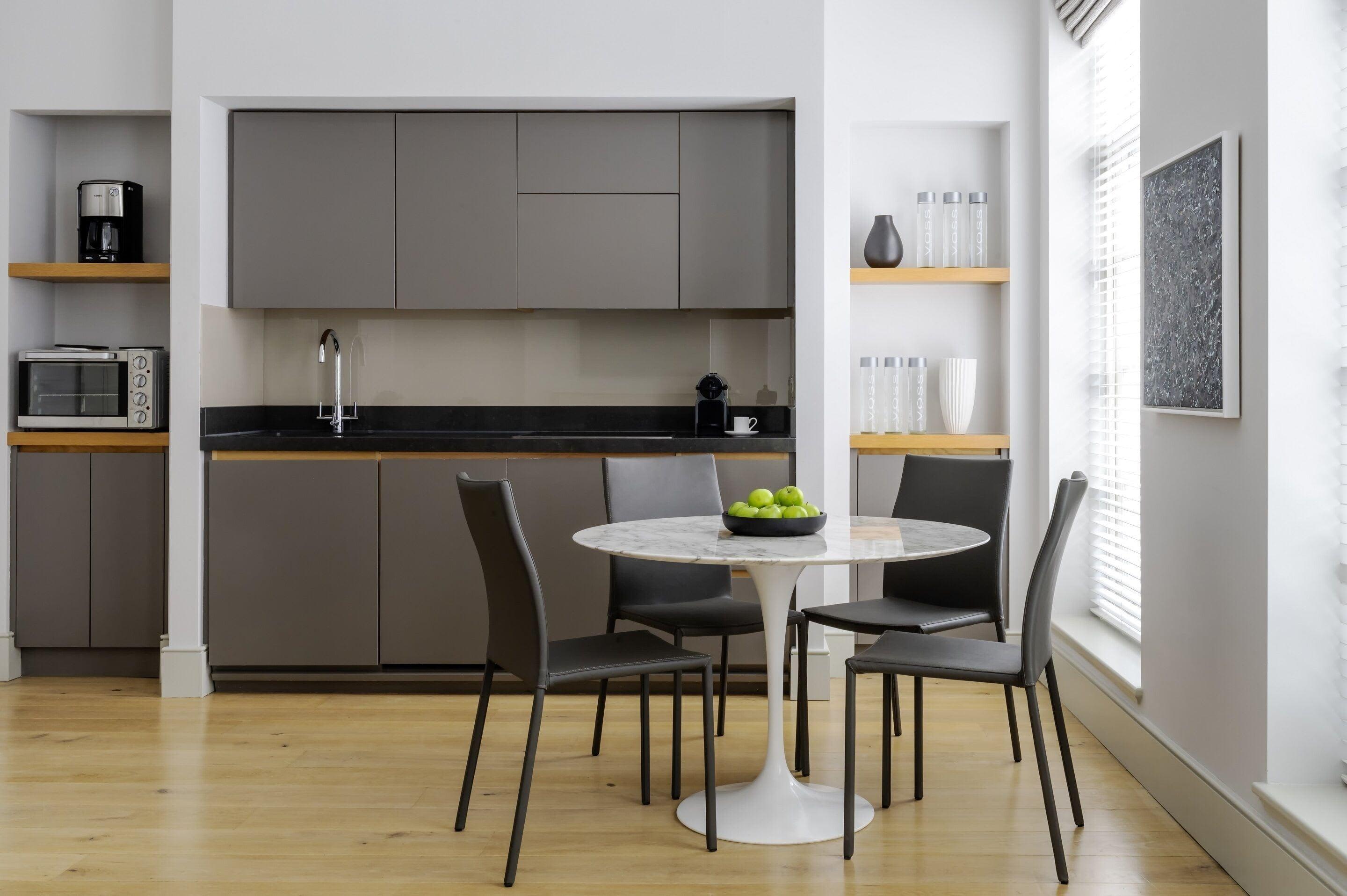 Open-plan kitchen and dining space with gray cabinetry, black countertop, round marble table, black chairs, and wooden shelving.
