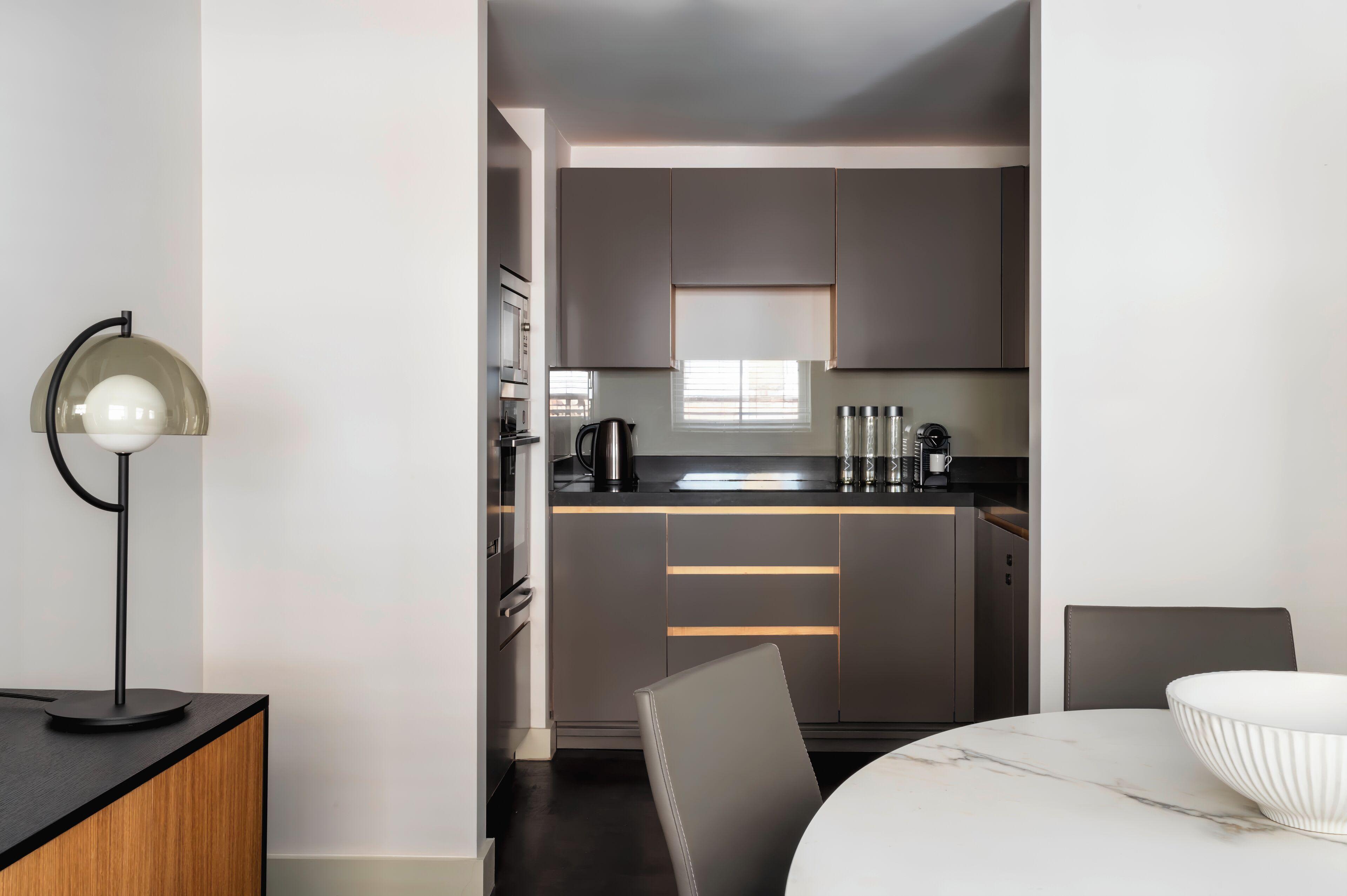 Studio bedroom with view into open-plan kitchenette; dark gray cabinets, black countertop, stainless steel appliances, and window with blinds.