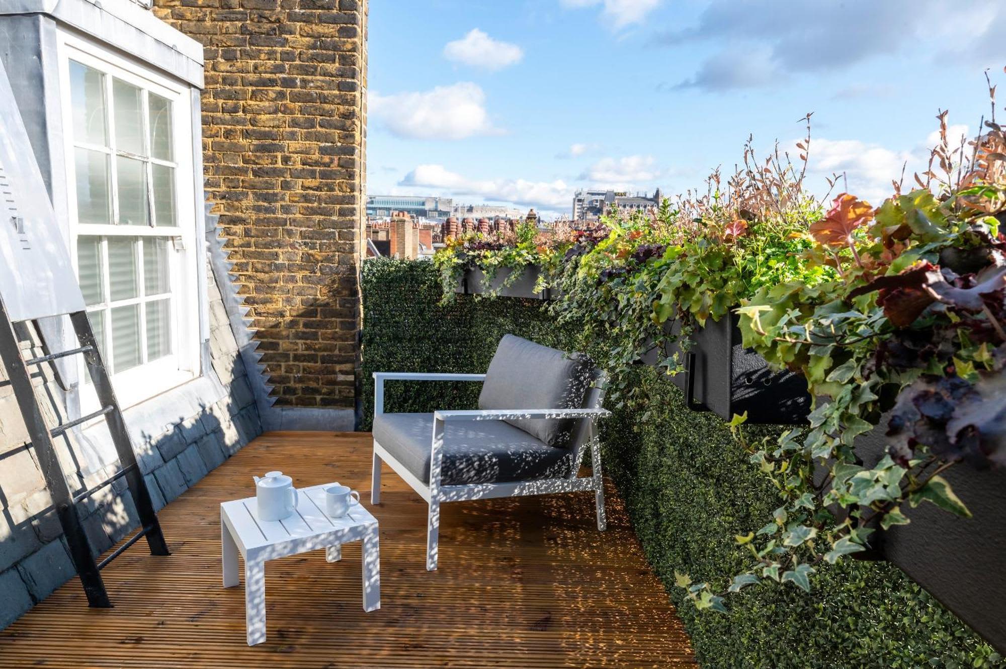 Rooftop terrace with wooden decking, white modern chair, small table, ivy-covered walls, and London cityscape in background.