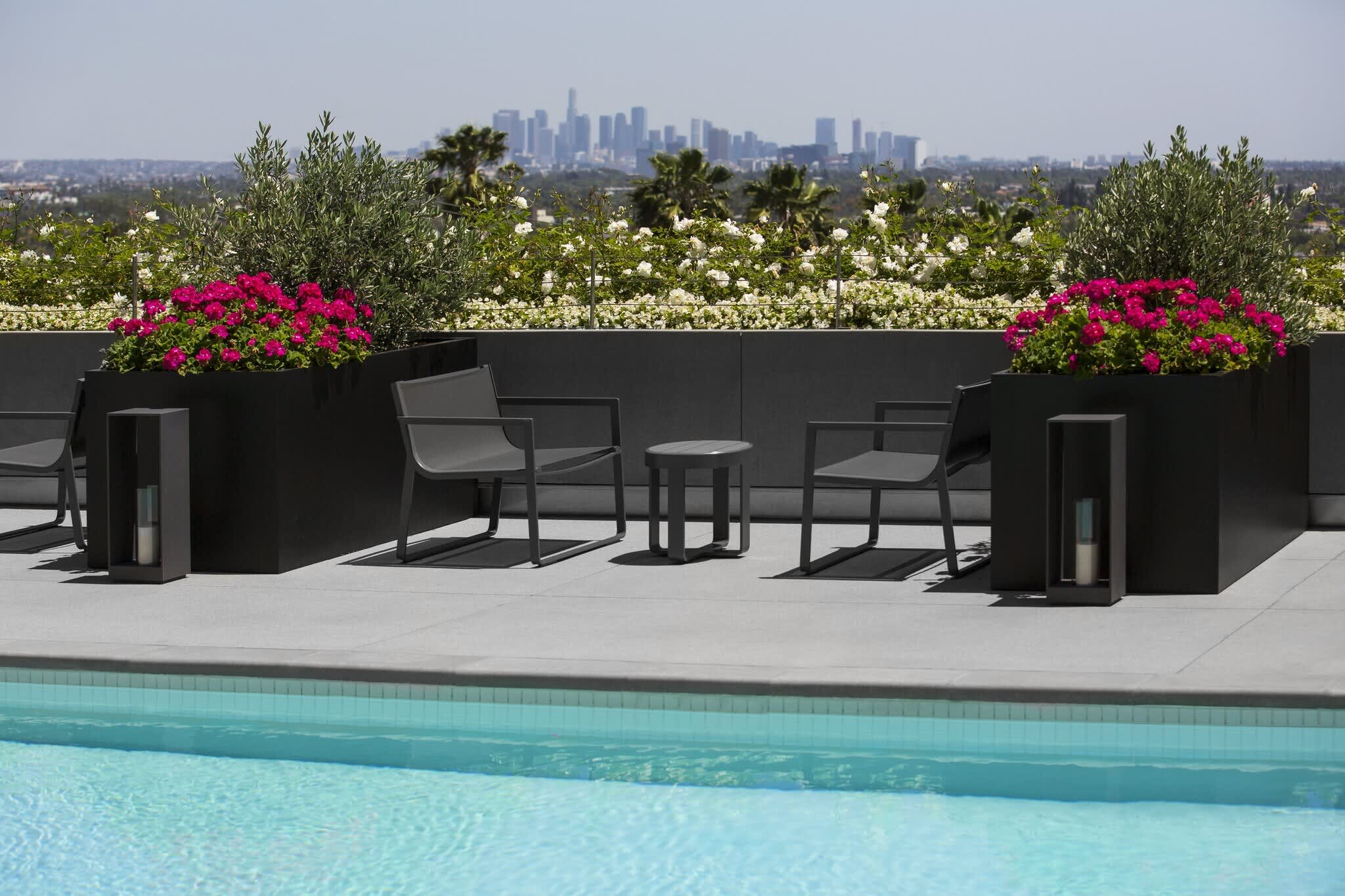 Rooftop pool deck with modern gray lounge chairs, black planters of magenta flowers, and downtown Los Angeles skyline in background.
