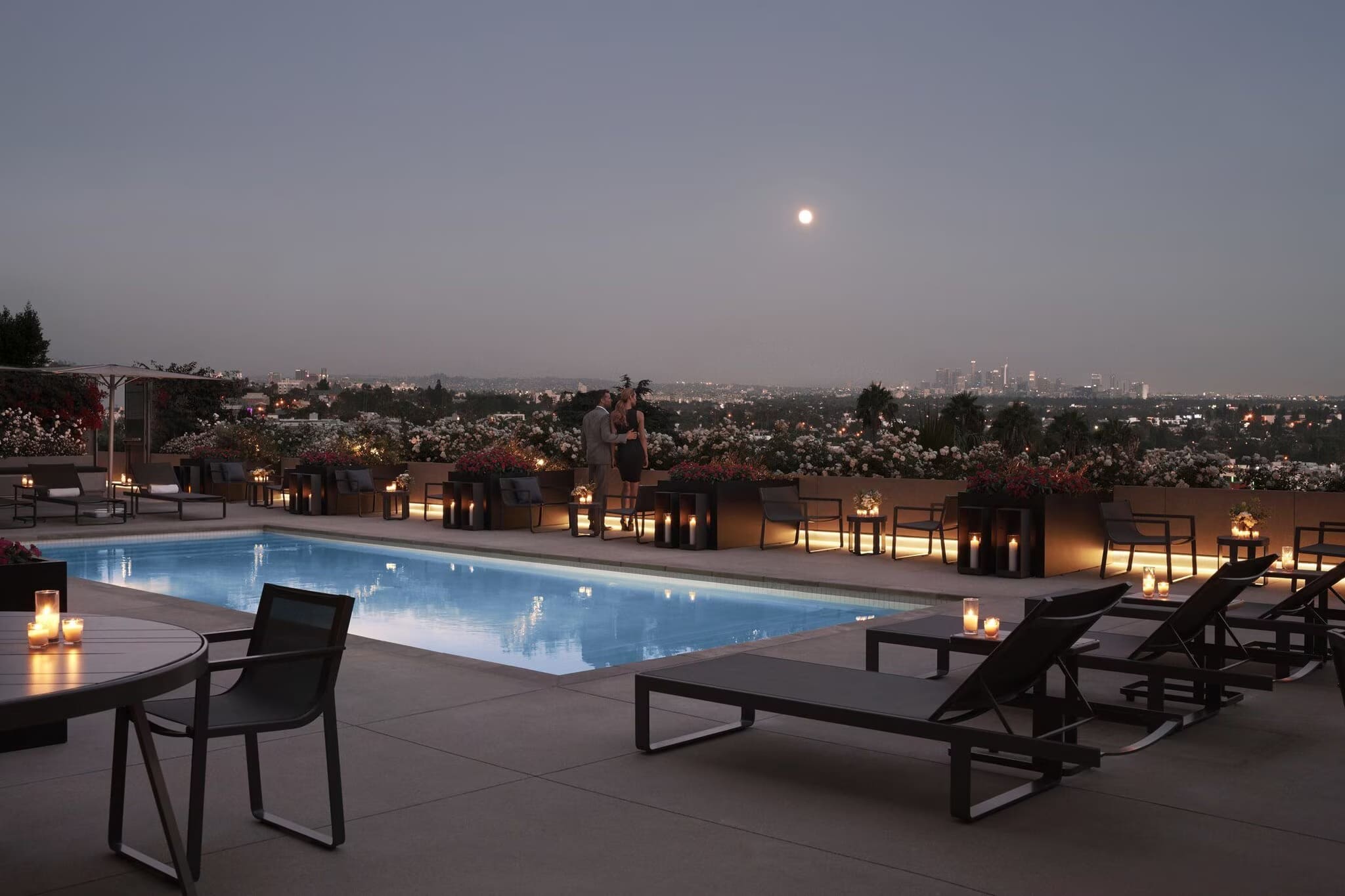 Rooftop pool at dusk with illuminated water, lounge chairs, lanterns, and West Hollywood cityscape under moonlit sky.