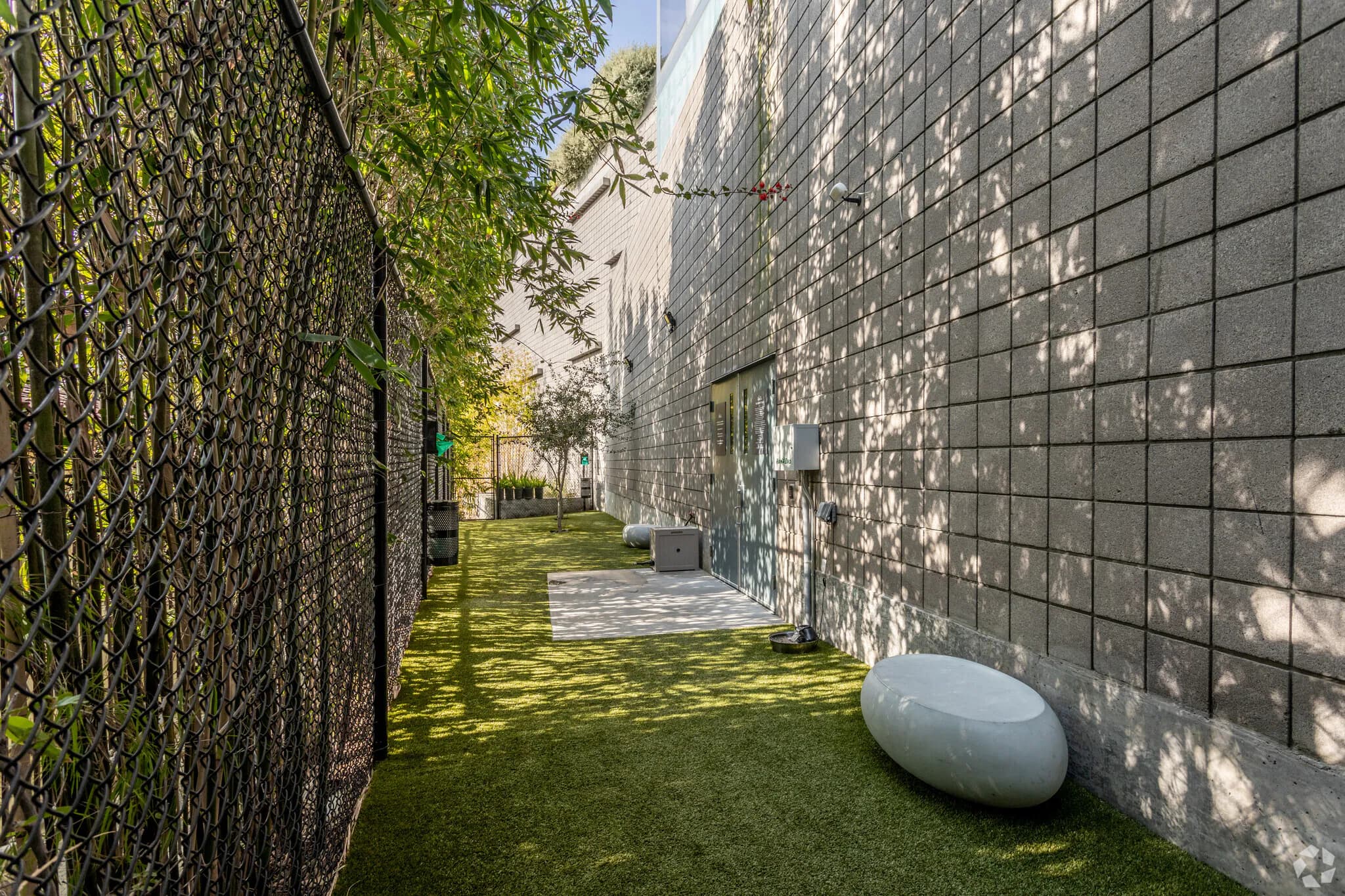 Narrow outdoor courtyard with synthetic grass, ivy-covered fence on left, gray tile building wall on right, white sculptural seating, dappled sunlight.