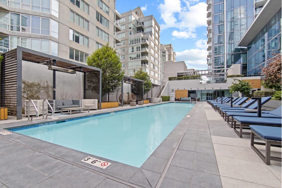 Outdoor swimming pool with blue lounge chairs, wooden pergola seating area, and surrounding residential towers under clear sky.