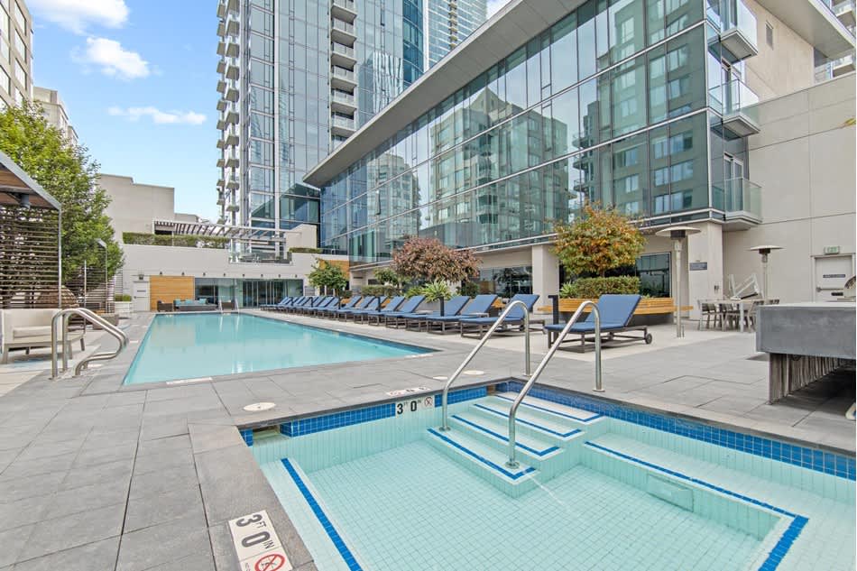 Outdoor pool deck with two pools, blue lounge chairs, spa, and surrounding high-rise residential buildings under clear sky.