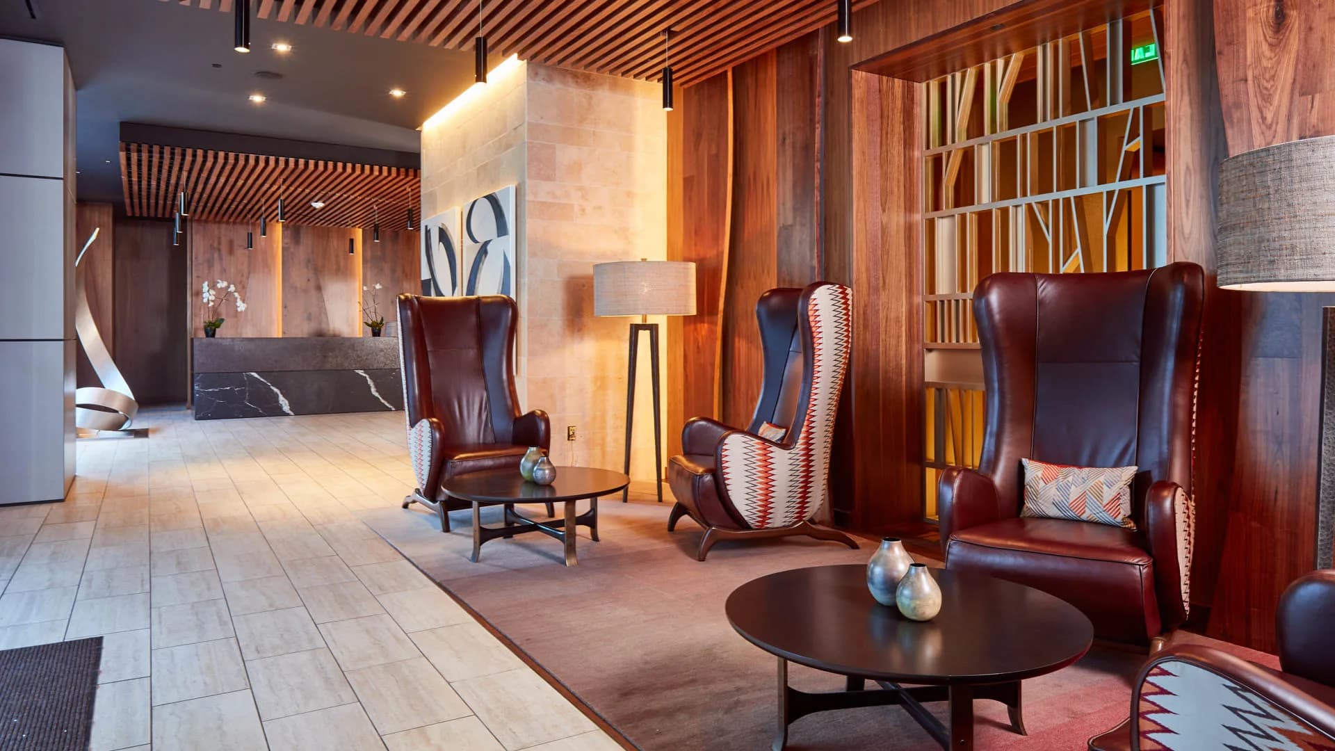 Modern apartment lobby with burgundy leather wingback chairs, wood slat ceiling, round black tables, and geometric patterned rug.