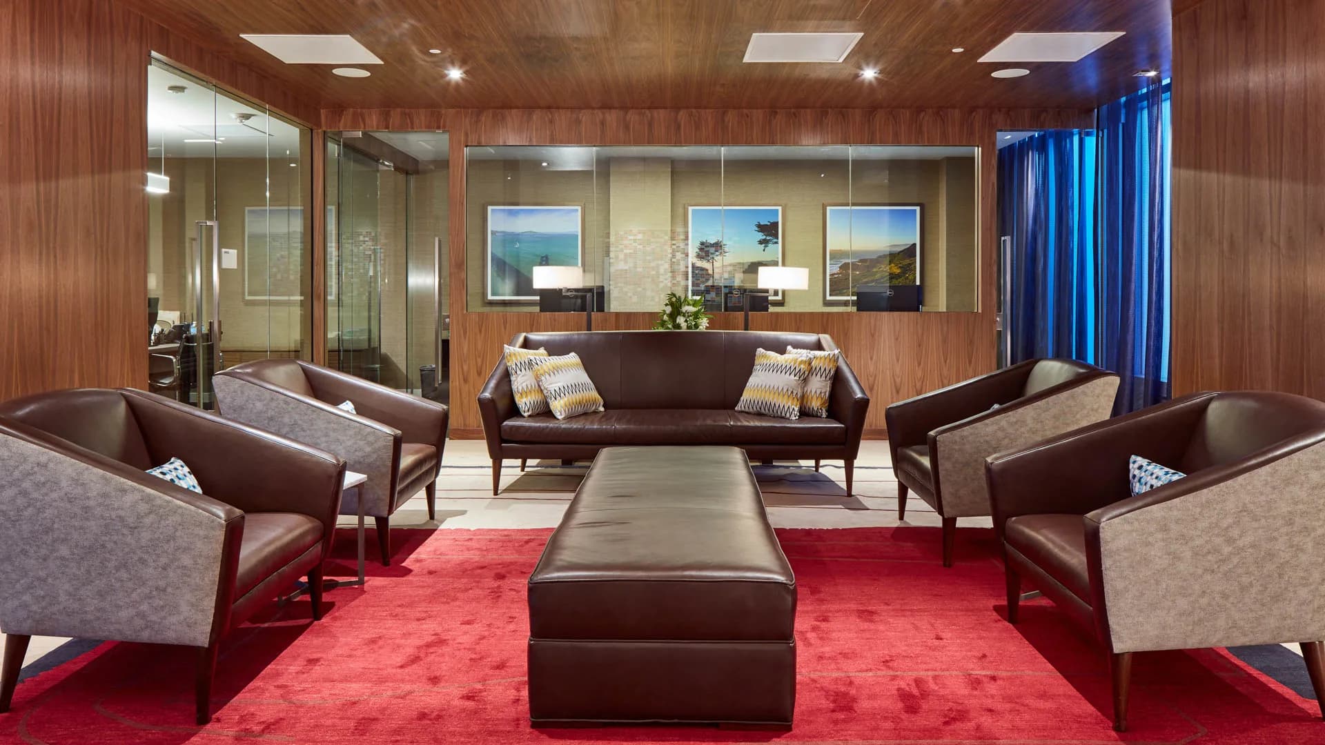 Modern lounge with brown leather sofa, upholstered armchairs, red carpet, wood-paneled walls, and framed landscape photos.