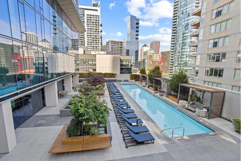 Rooftop pool deck with rectangular swimming pool, blue lounge chairs, planters with greenery, and surrounding high-rise buildings under blue sky.