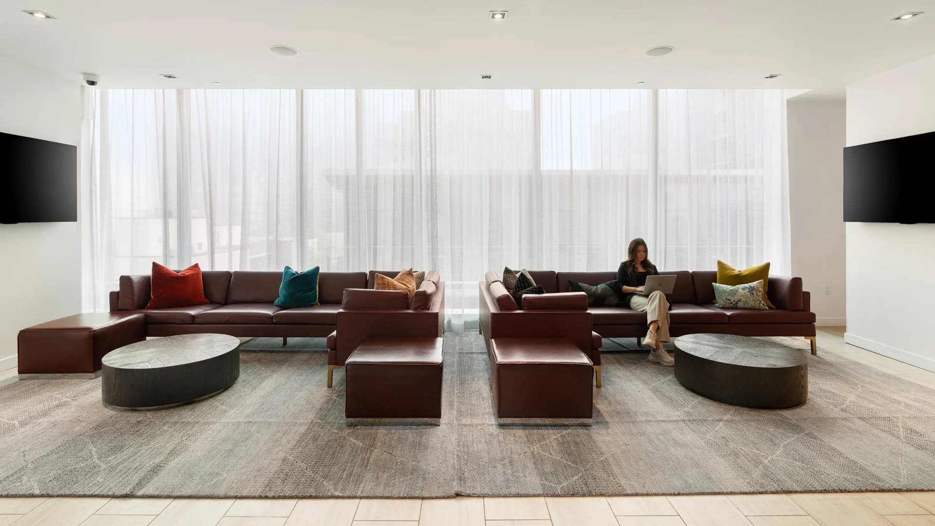 Modern lounge with burgundy sectional sofas, colorful accent pillows, oval marble tables, and floor-to-ceiling windows with sheer curtains.